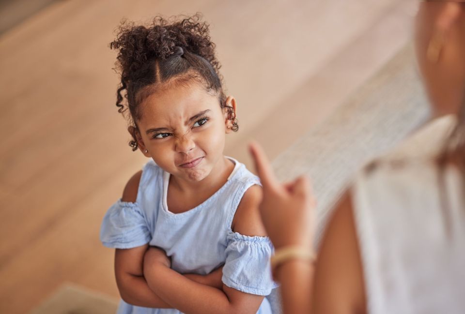 What is a positive behavior support plan? Why your child might need one What is a positive behavior support plan? Why your child needs one. A young black girl in a light blue ruffled top is making a displeased face with furrowed brows while standing indoors. She is crossing her arms and pointing upwards with one finger, expressing disapproval or making a point during a discussion with an adult, whose presence is implied but not shown in the image. The child's curly hair is styled in small buns.