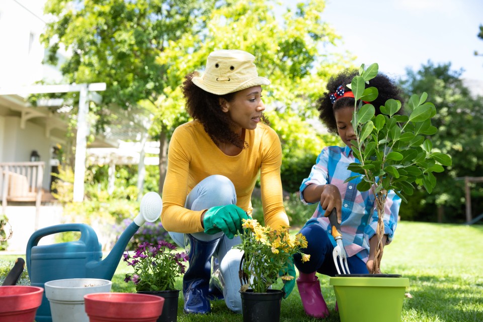 Practical tips for creating a stylish garden on any budget A black mother wearing a sun hat and green gloves kneels on the lawn beside her young daughter, who uses a hand fork to pot a small tree, surrounded by colorful plant pots and a watering can in a sunny backyard.