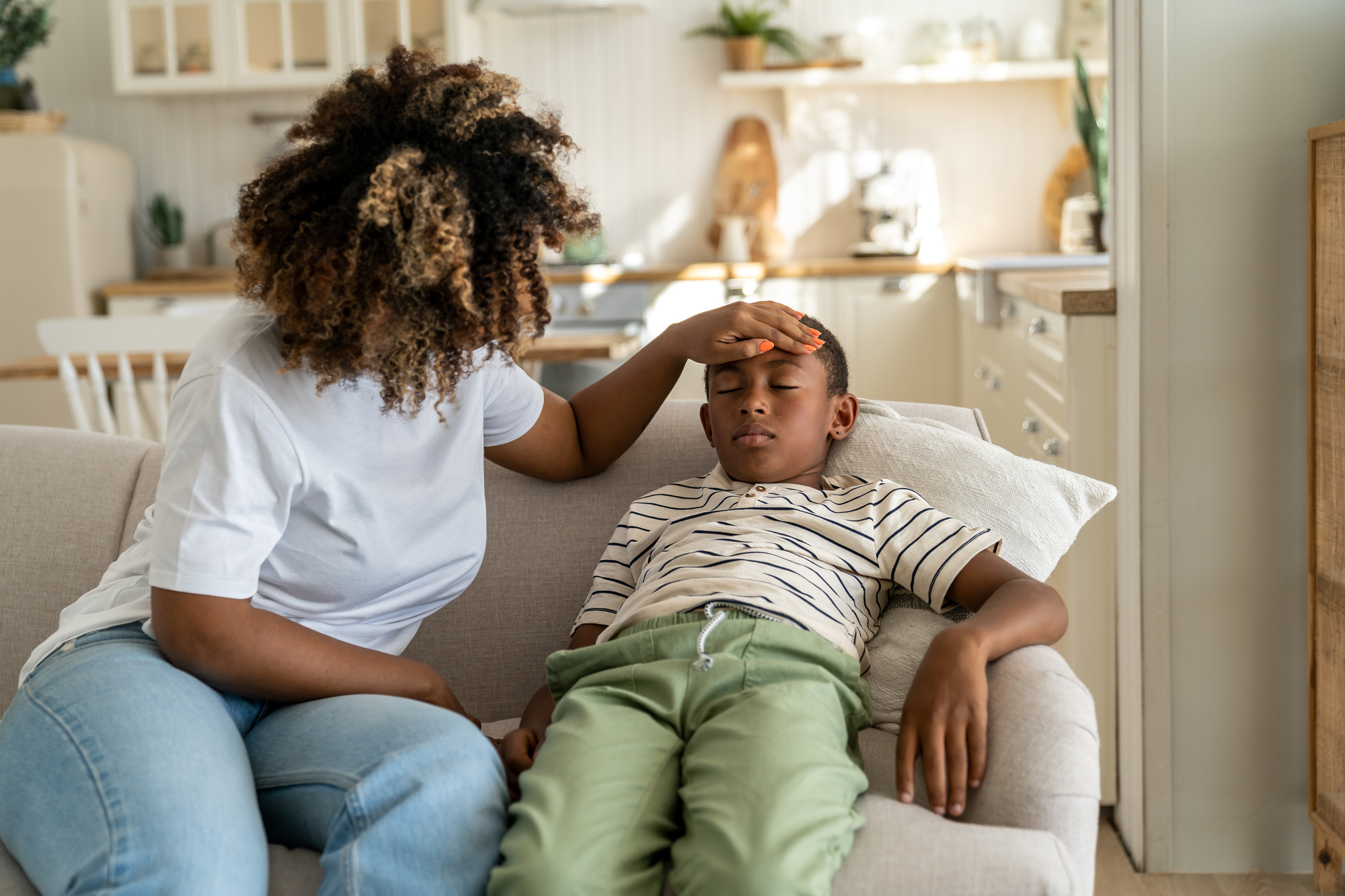 Surviving flu season: 5 essential tips for protecting your family Fever in kids. African american mother touching forehead of sick little boy son at home, caring parent mom checking childs temperature with hand. Schoolboy lying on sofa feeling unwell and ill during flu season.