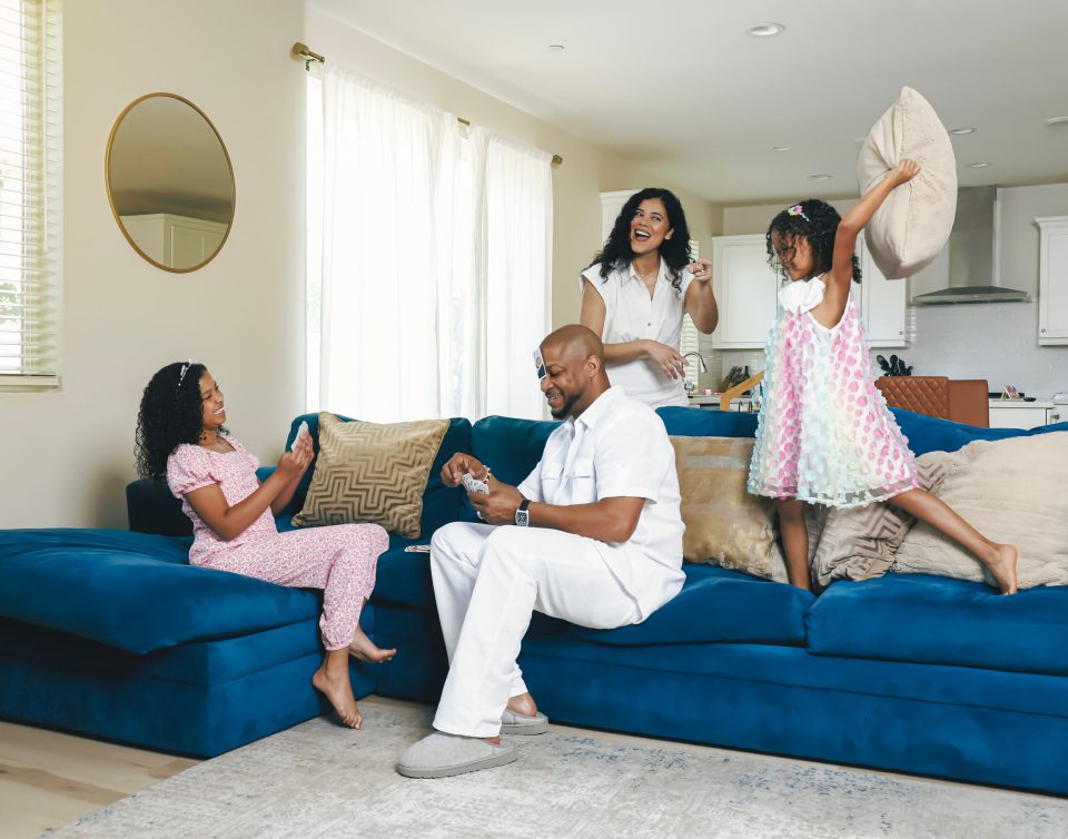 Finesse mitchell sits on a blue couch playing cards with his eldest daughter elle kate to his left, while his wife adris debarge smiles behind and their younger daughter eva kristian raises a pillow overhead.