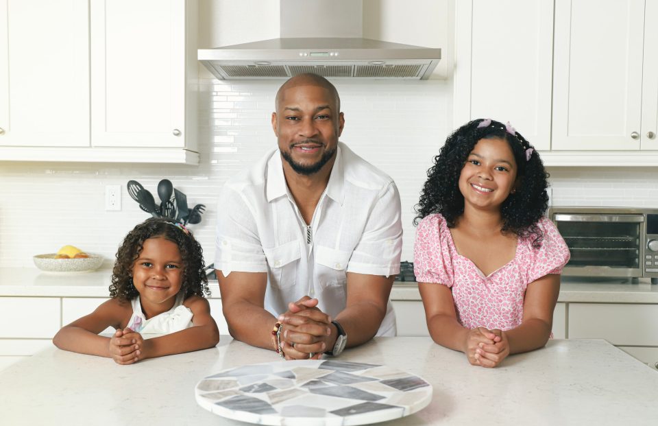 Finesse mitchell leans on a white kitchen countertop between his daughters—eva kristian on his left and elle kate on his right—all three smiling warmly into the camera.