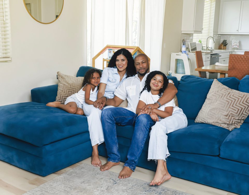 Finesse mitchell and his wife adris debarge sit on a blue sectional with their daughters, eva kristian on adris’s lap and elle kate leaning against finesse’s leg, as a family portrait in their living room.