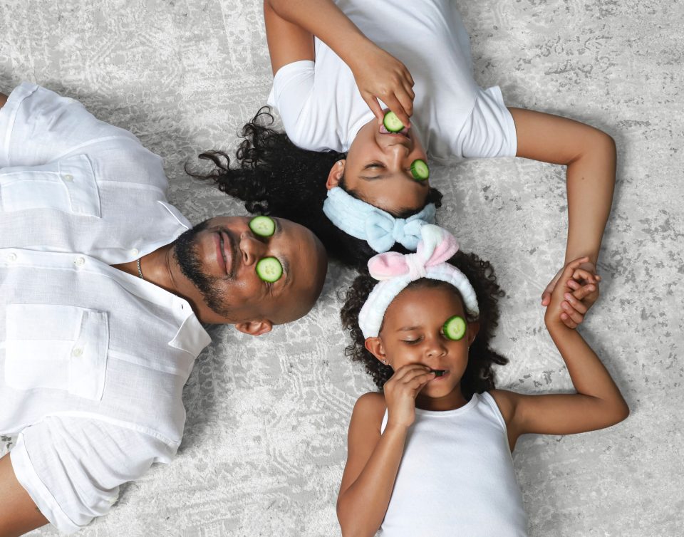 Finesse mitchell lies on a light gray rug holding hands with his daughters, elle kate and eva kristian, each wearing a cucumber slice over one eye for a playful at-home spa moment.