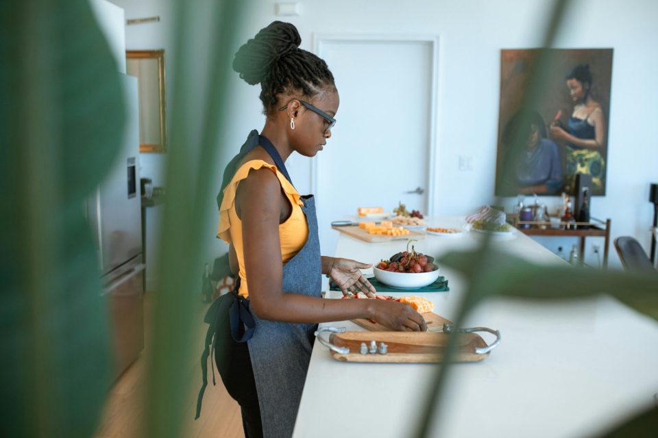 7 essential tips for running a home-based catering service Black woman preparing food in a modern kitchen while running a home-based catering service, demonstrating one of the 7 essential tips for success for an article titled, "7 essential tips for running a home-based catering service. "
