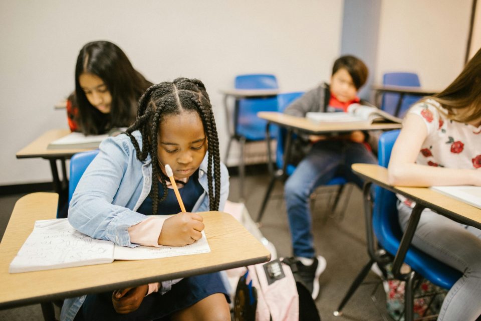 How school access control systems keep your child safe: a parent’s guide A young black girl writing in a workbook at her school desk, surrounded by classmates in a classroom setting—illustrating the importance of student safety and focused learning supported by school access control systems.