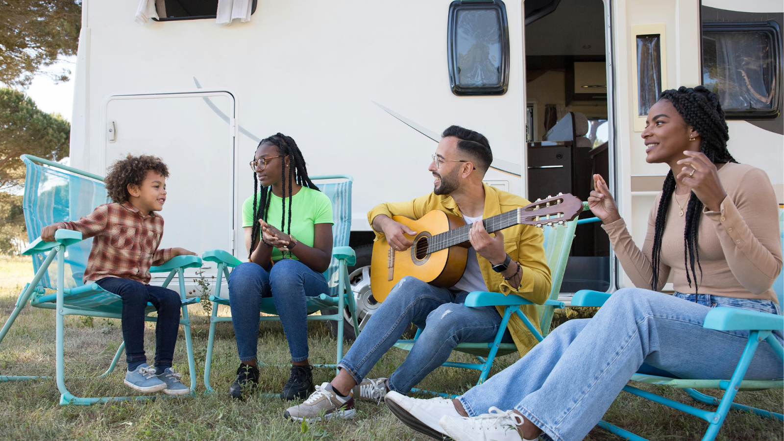 7 smart tips for traveling with kids this summer (without losing your mind) A family of four sits in folding lawn chairs outside a camper van; a young black boy, a black teenage girl, a father playing guitar, and a black mother smiling and singing along.