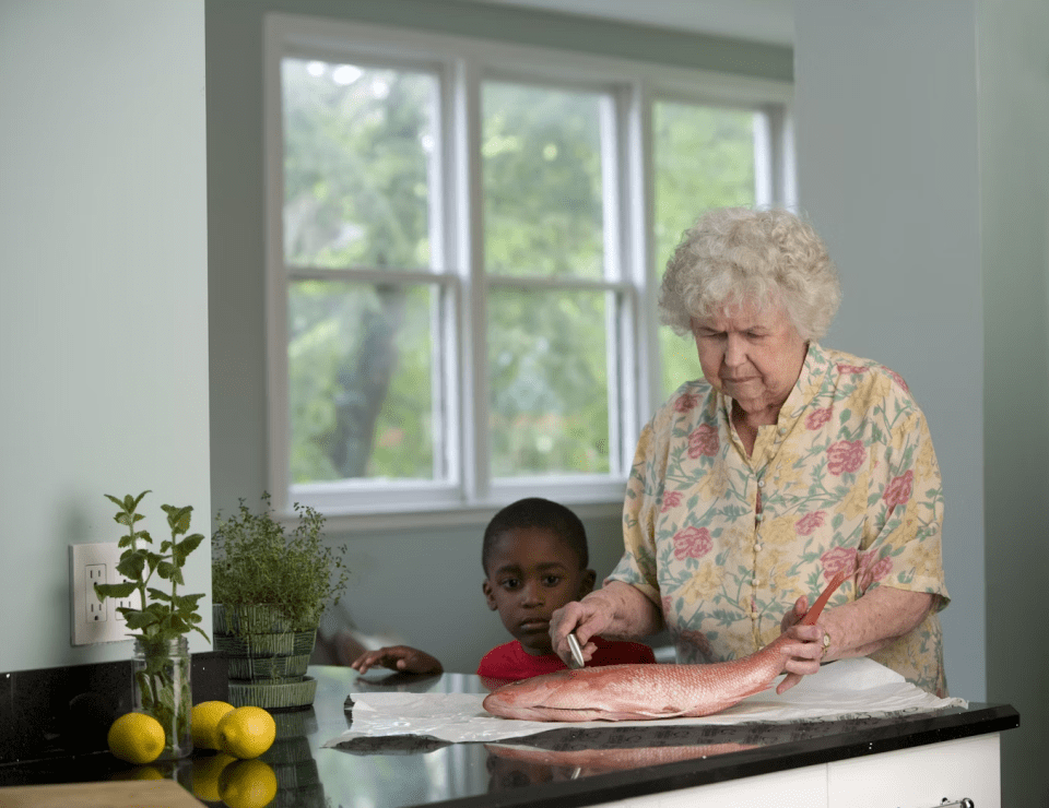 Could assisted care be the right choice for your family? An elderly woman prepares a whole fish at the kitchen counter while a young black boy watches attentively, symbolizing intergenerational caregiving and early signs of when assisted care may be needed.